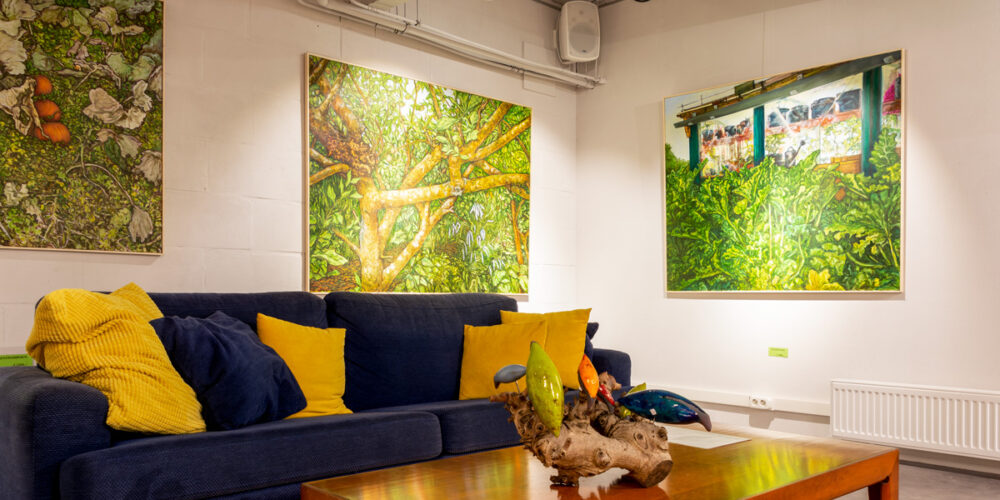 drie schilderijen met veel natuurgroen en een zitbank met kussens in de expositieruimte van Het Gele Huis