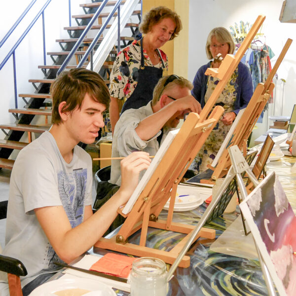 een jongen en een man achter een schilderezel tijdens een schilderworkshop. Docenten kijken toe